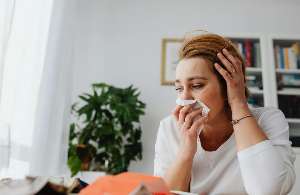 A woman blowing her nose at a table, symbolizing the impact of fall allergies, highlighting the need for an effective HVAC system.