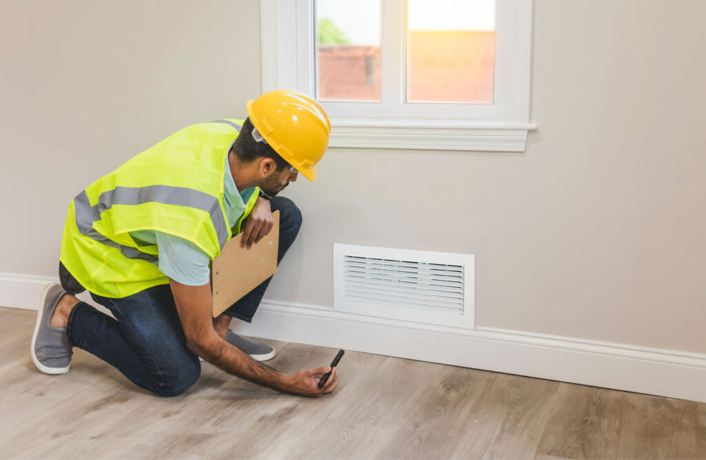 A technician in a hard hat and safety vest kneeling while inspecting an HVAC unit during fall maintenance.