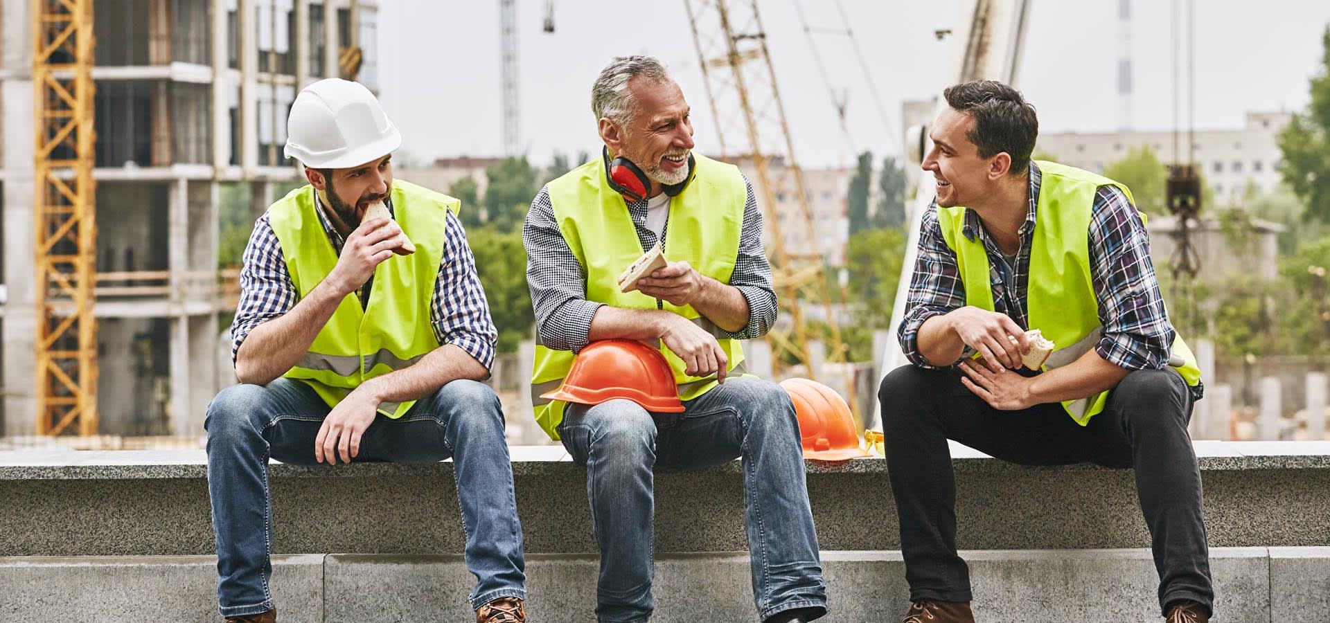Three smile and construction workers converse over a lunch of sandwiches on the jobsite