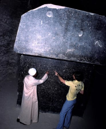 Two men inside an egyptian pyramid looking at inscriptions on a black slab