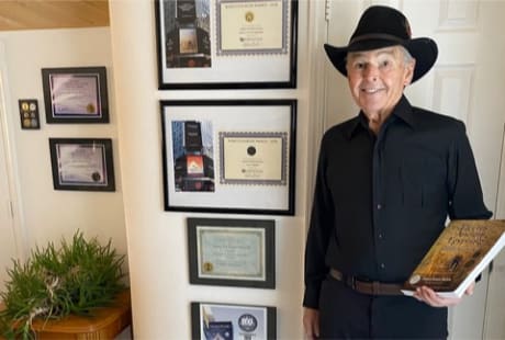 James E. Brown holding one of his books and standing next to a display of awards and certifications.