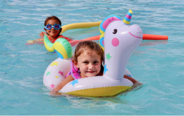 Two toddlers playing in the mineral water pool. One in the front using a unicorn inflatable ring and the ther one floating on two pool noodles.