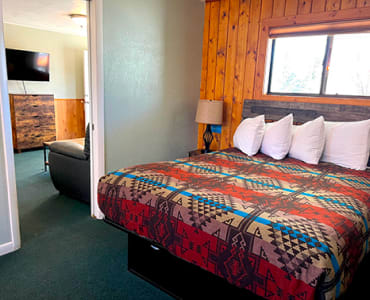 A suite room with wood-paneled walls, it showcase a queen bed, and a living room visible through the door with a TV and a couch.